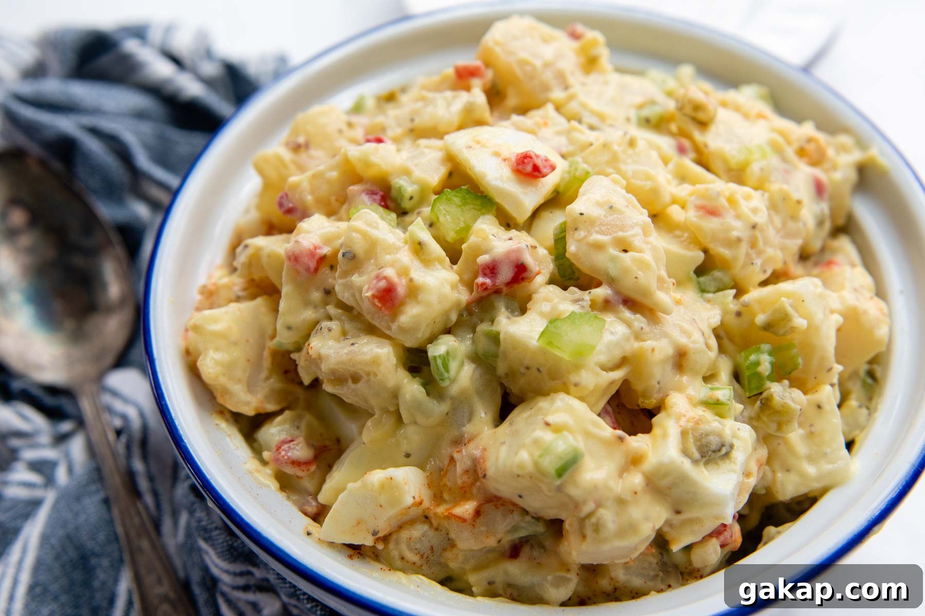 a horizontal view of a bowl of potato salad with a blue linen and serving spoon next to it.