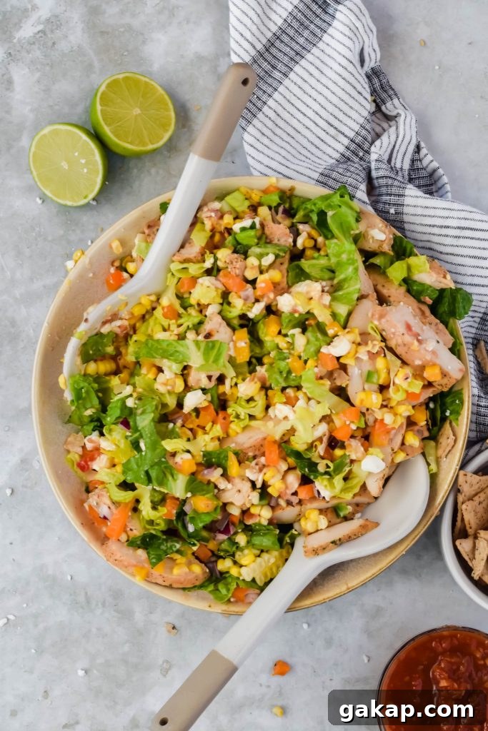 Tossed Mexican caesar salad in cream bowl with salad tongs in bowl on slate background beside lime wedges.