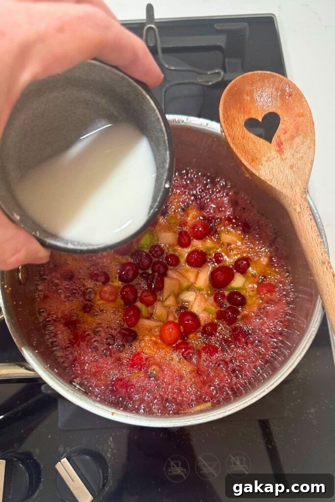 Apple Cranberry Sourdough Hand Pies Recipe for Flaky Pastries 8 A bowl of corn starch and water being poured into the saucepan of apple and cranberry mixture that has simmered down on the stove top.
