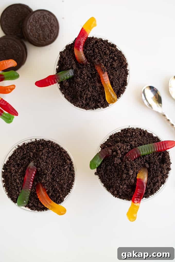 No-Bake Dirt Cake Cups Recipe — Creamy Chocolate Pudding Cups 5 overhead image of cups full of crushed cookies and gummy worms on a white surface