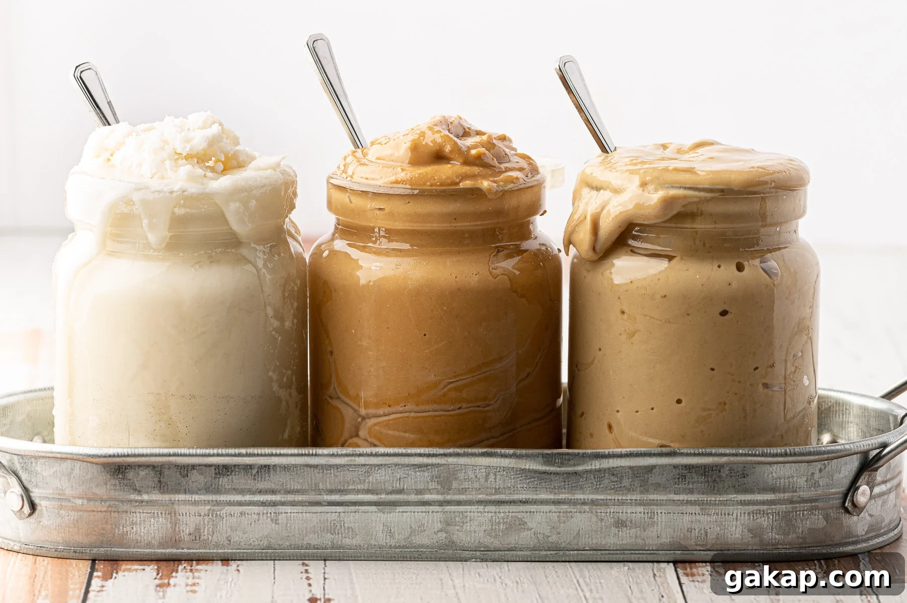 Top Nut Butters for Low-Carb Baking: Almond, Peanut & More 2 A shallow rustic metal tray with 3 jars of different nut butters spilling out over the jar rims.