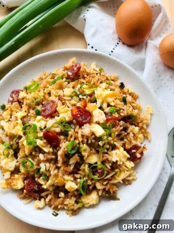 A full plate of Chinese sauage fried rice. Raw spring onions, eggs and spoon in the background sat on top of a tea towel