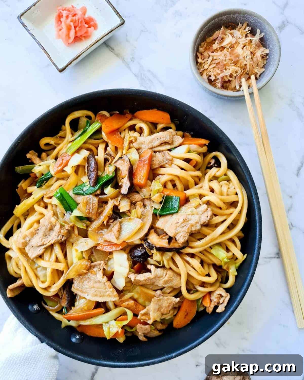 Stir fried udon in a black pan with bonito flakes and pickled ginger in individual dishes on the side.