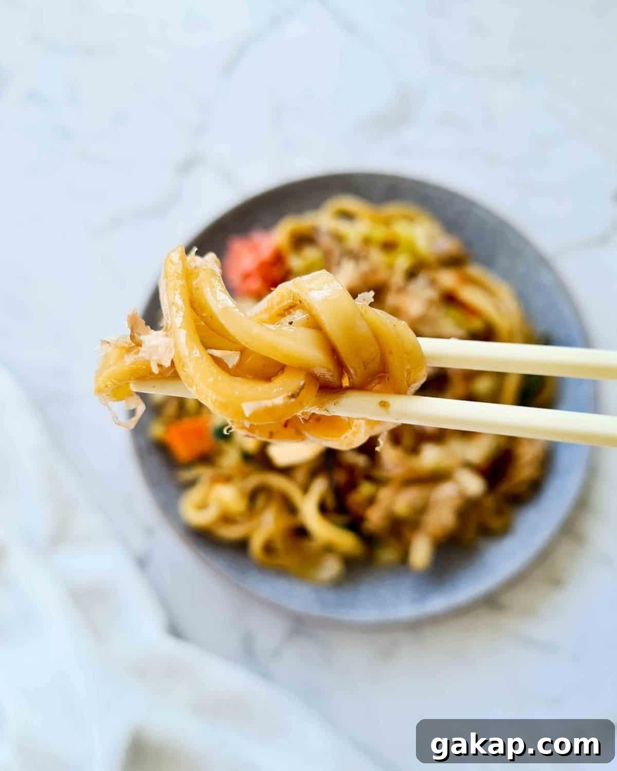close up mouthful of udon held up by a pair of chopsticks. Blurred out plate of noodles in the background