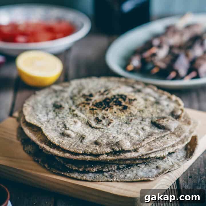 a stack of buckwheat pita breads on a wooden board with souvlaki sticks and tomatoes in the background.