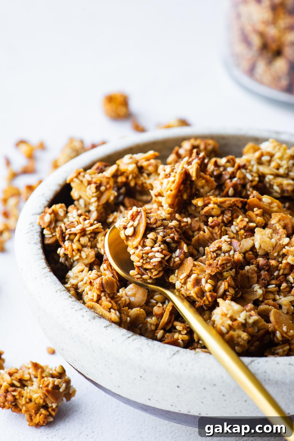Tahini Sesame Granola with Honey and Orange Zest 3 Close-up of tahini granola with sesame seeds in a ceramic bowl with a jar in the background and a small gold spoon.