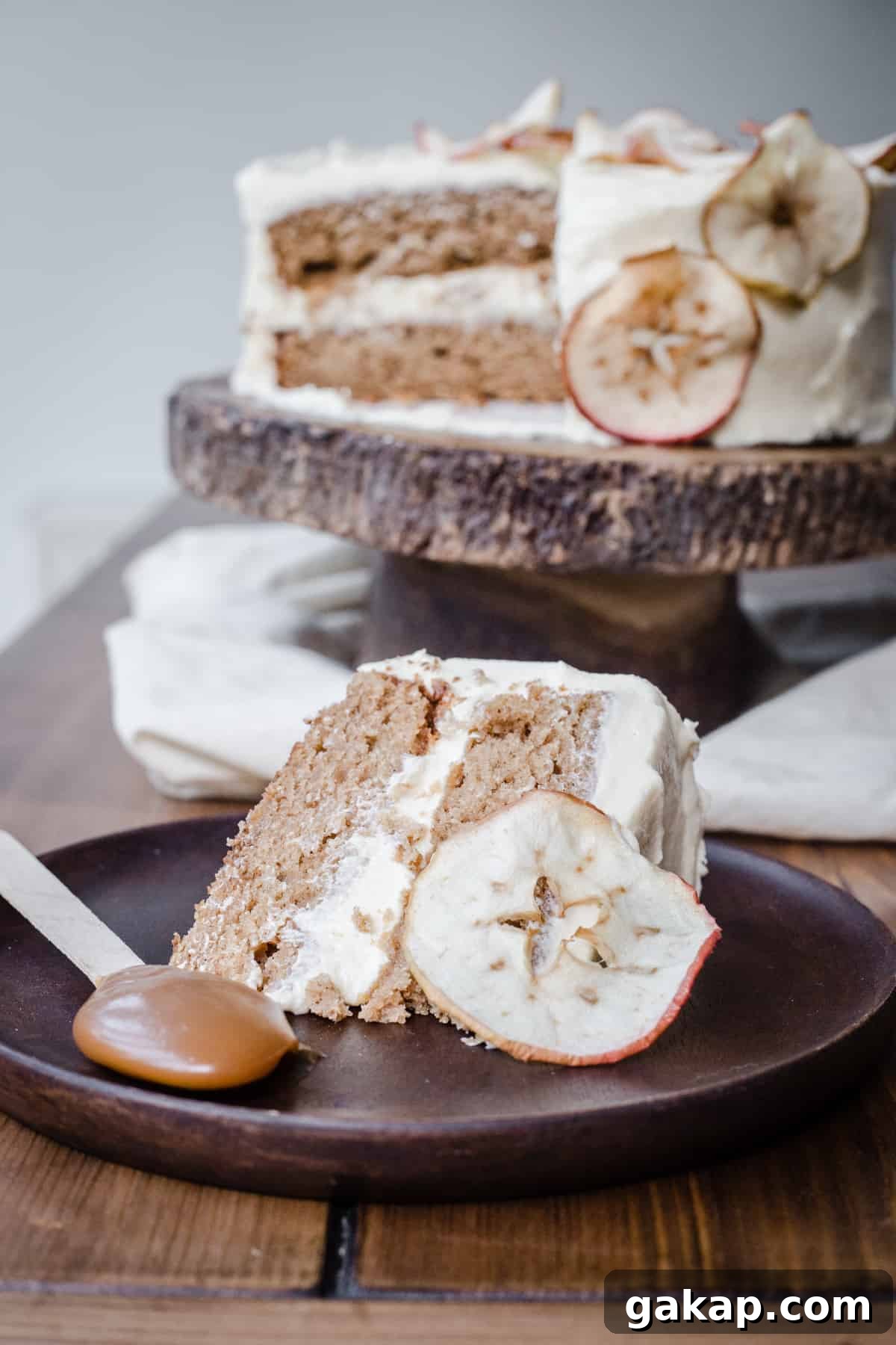 A slice of Cinnamon Apple Cake with Salted Caramel Cream Cheese Buttercream on a wooden plate with an apple slice and a spoon of caramel, infront of the whole cake