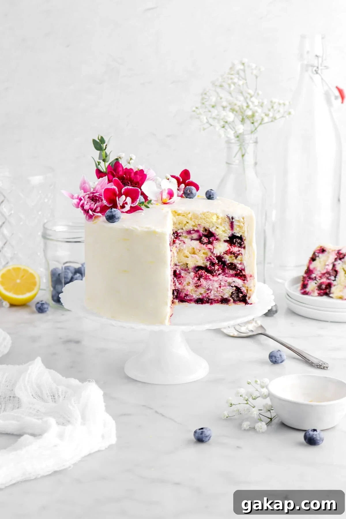 Lemon Blueberry Layer Cake with Zesty Lemon Buttercream 2 pulled back front shot of frosted lemon blueberry layer cake with flowers on top and slice of cake on stack of three plates behind, fresh blueberries in a glass jar, and a lemon half behind with more blueberries round, a small white bowl, and a white cheesecloth on marble surface.