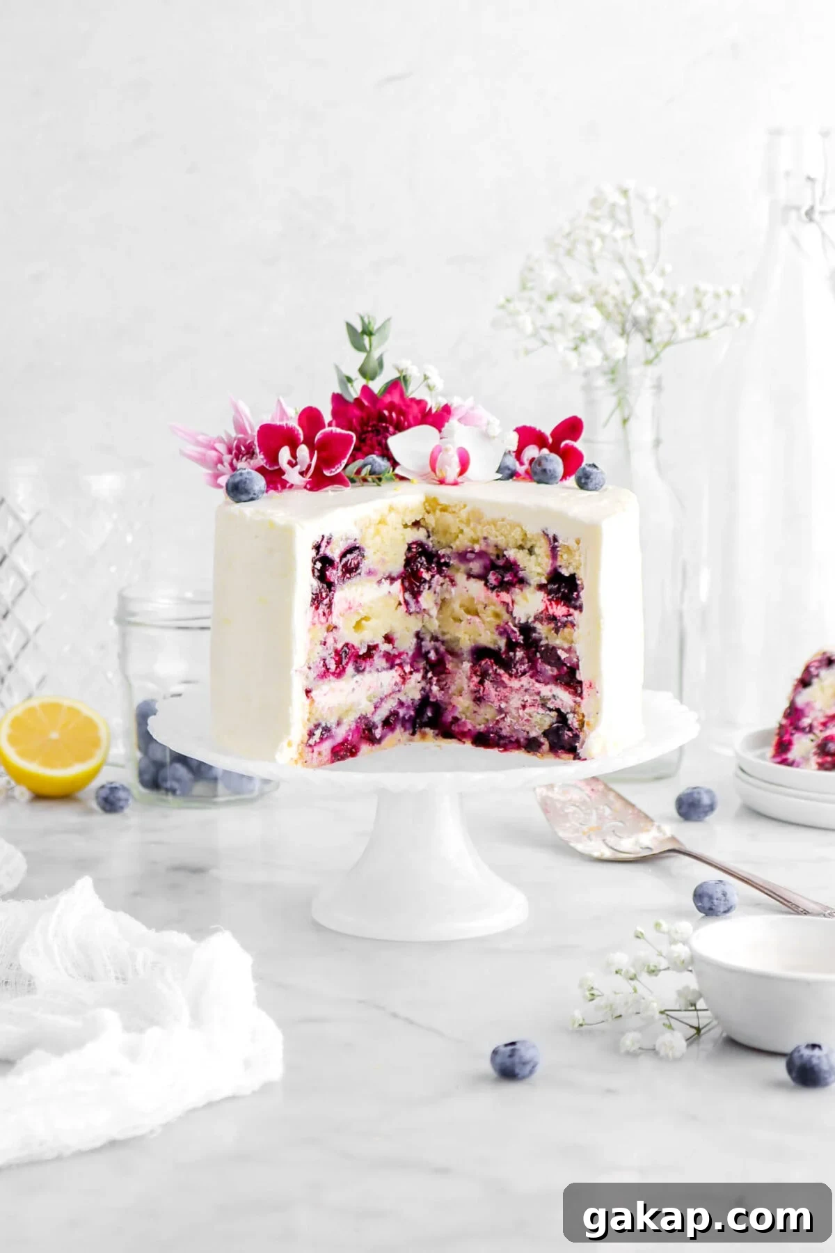Lemon Blueberry Layer Cake with Zesty Lemon Buttercream 3 cut lemon blueberry cake on white cake stand with flowers on top, and cake slice behind, a cake knife beside, and blueberries around, and a lemon half behind and empty glasses on marble surface.