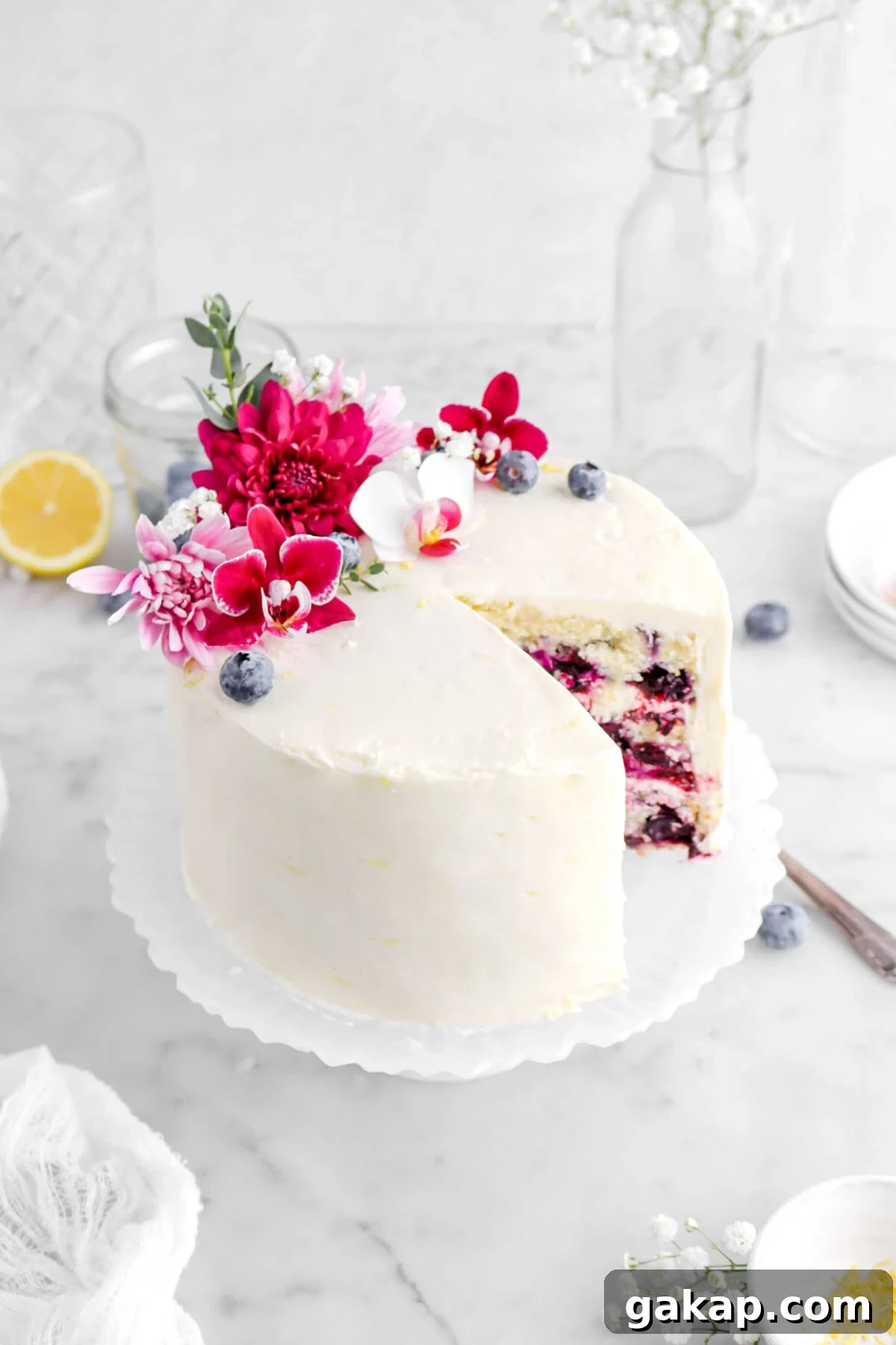 Lemon Blueberry Layer Cake with Zesty Lemon Buttercream 4 angled close up of lemon blueberry layer cake decorated with flowers on top with lemon half and empty glass behind on marble surface.