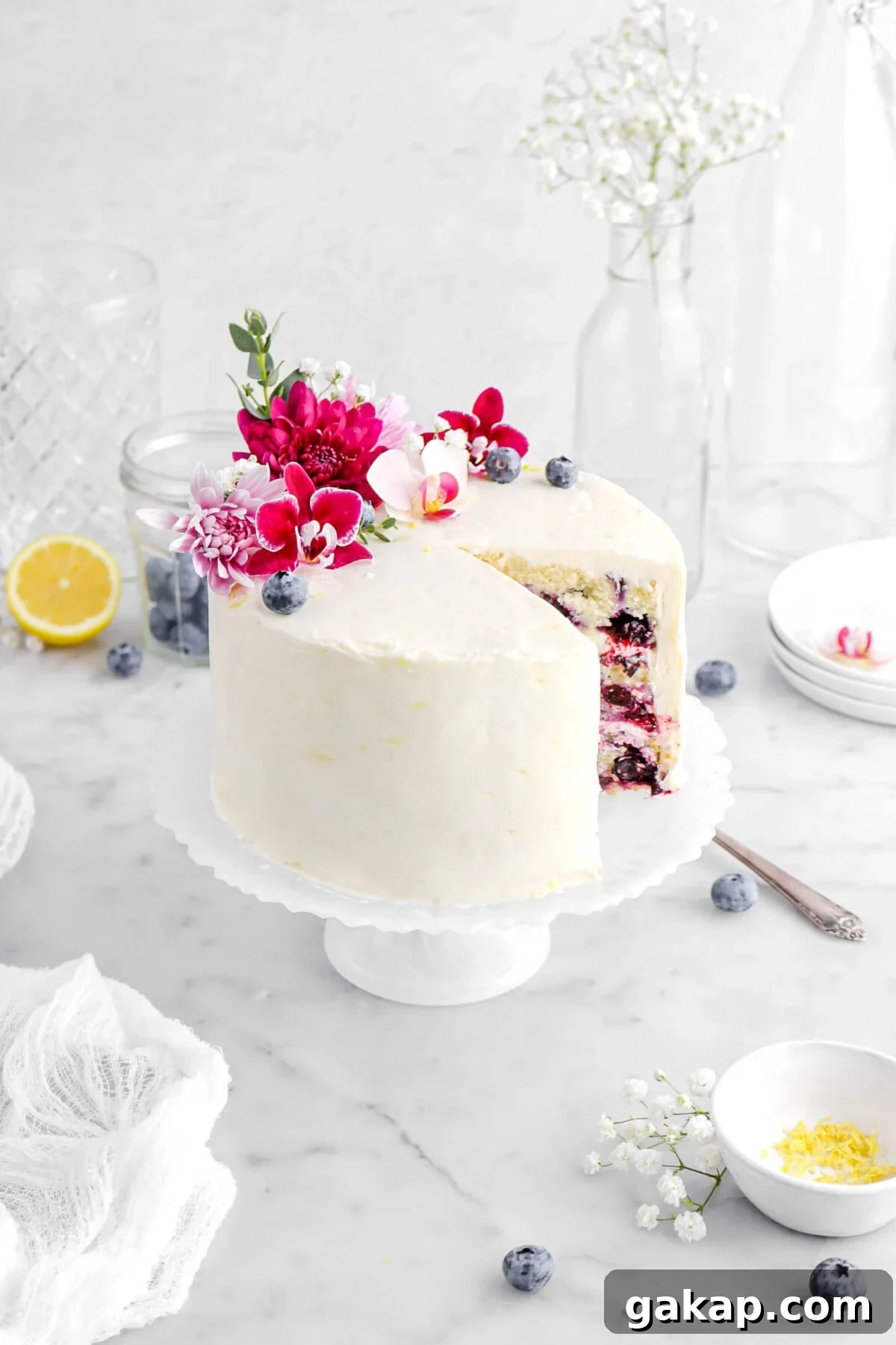Lemon Blueberry Layer Cake with Zesty Lemon Buttercream 15 lemon blueberry cake with slice missing on white cake stand with flowers and blueberries on top, a bowl of lemon zest beside, white flowers around, a white cheesecloth beside, and empty glasses behind.