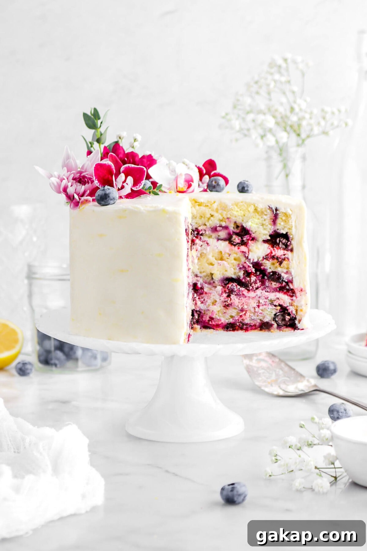 Lemon Blueberry Layer Cake with Zesty Lemon Buttercream 16 close up of cut lemon blueberry cake with blueberries and flowers on top on a white cake stand with blueberries and white flowers around on marble surface.