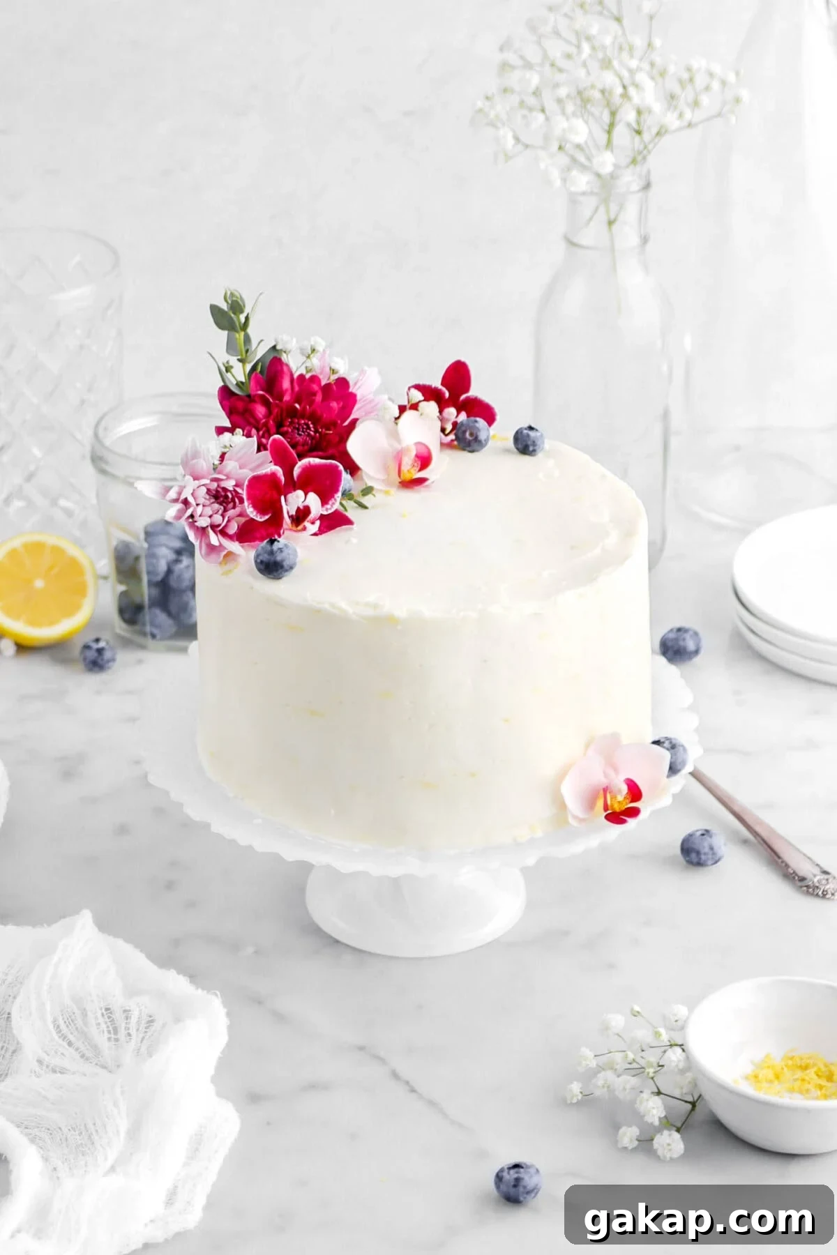 Lemon Blueberry Layer Cake with Zesty Lemon Buttercream 5 angled close up cake on white cake stand decorated with pink flowers and blueberries, with white flowers around and more blueberries on marble surface.