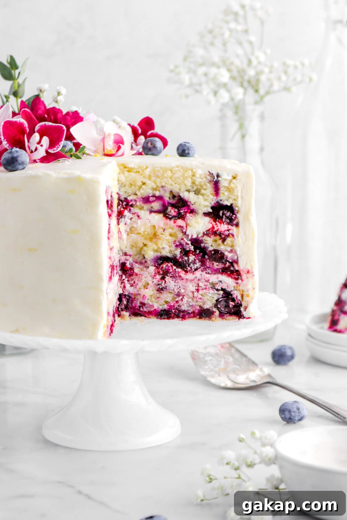 Lemon Blueberry Layer Cake with Zesty Lemon Buttercream 6 cropped close up of sliced lemon blueberry cake on white cake stand and pink flowers on top with blueberries beside and cake knife.
