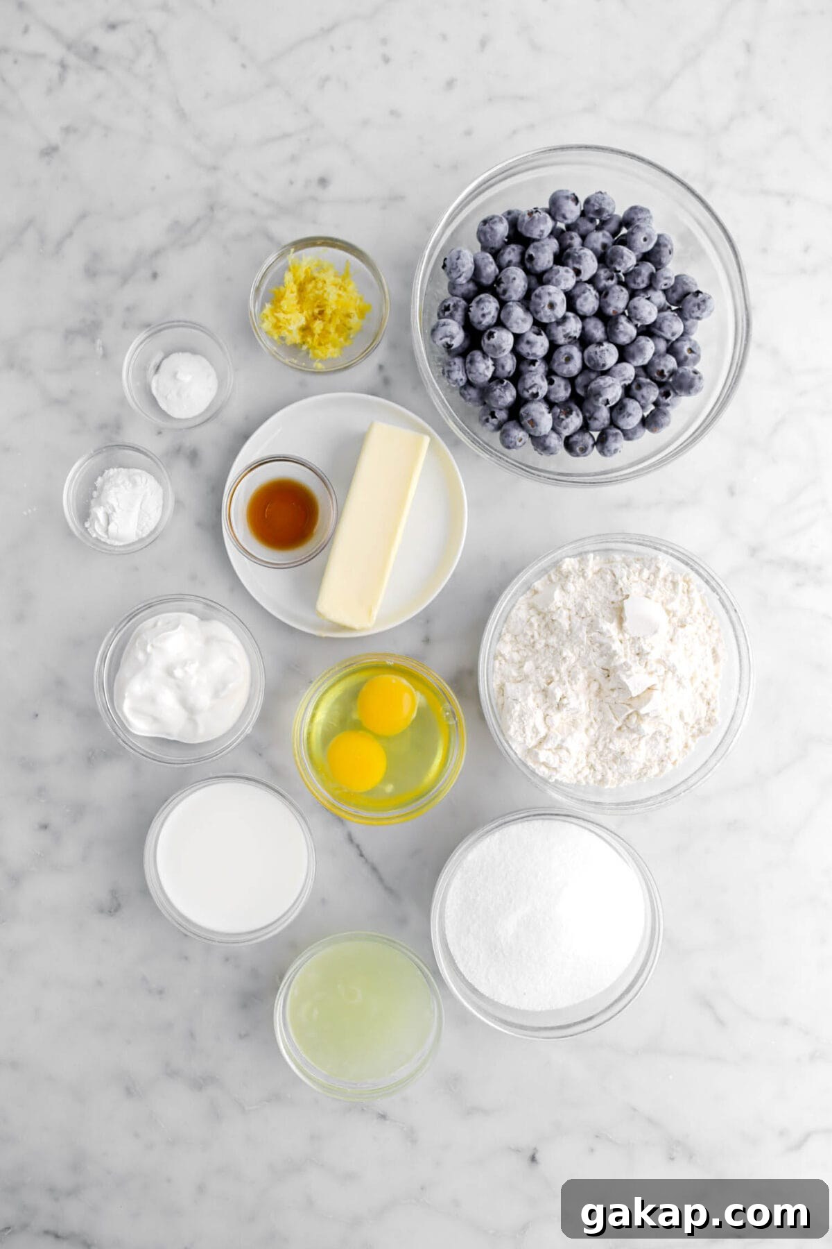 Lemon Blueberry Layer Cake with Zesty Lemon Buttercream 7 lemon zest, baking soda, baking powder, blueberries, vanilla, stick of butter, flour, eggs, sour cream, milk, sugar, and lemon juice on marble surface.