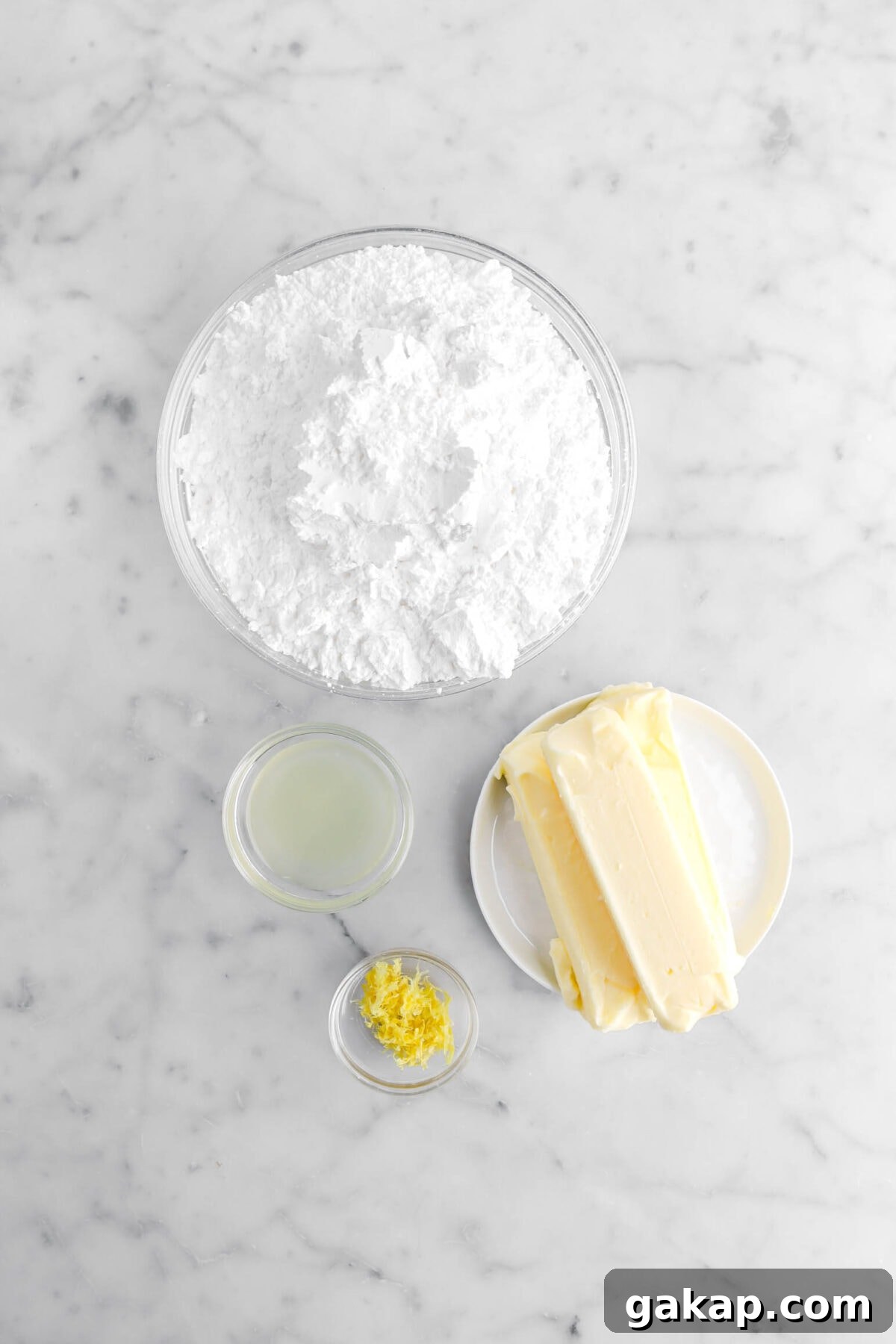 Lemon Blueberry Layer Cake with Zesty Lemon Buttercream 8 powdered sugar, butter, lemon juice, and lemon zest on marble surface.