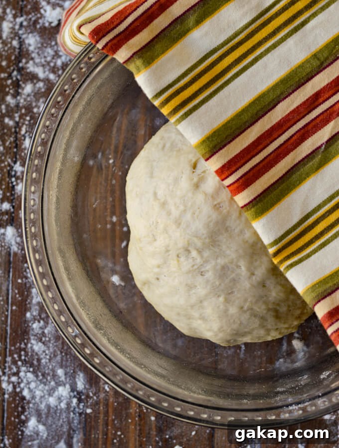 Crispy-Chewy Homemade Pizza Dough Recipe for Perfect Crust 6 A glass bowl with a ball of pizza dough ready to rise. There is a striped napkin over have the bowl and flour sprinkled around on the surface.