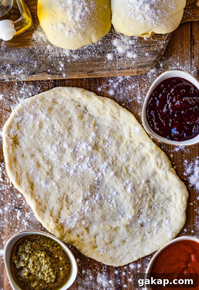 Crispy-Chewy Homemade Pizza Dough Recipe for Perfect Crust 7 A rolled out pizza dough with two more balls of dough sitting on a wooden board with flour sprinkled all around. Theres a jar of olive oil and three bowls of different sauces beside the dough.