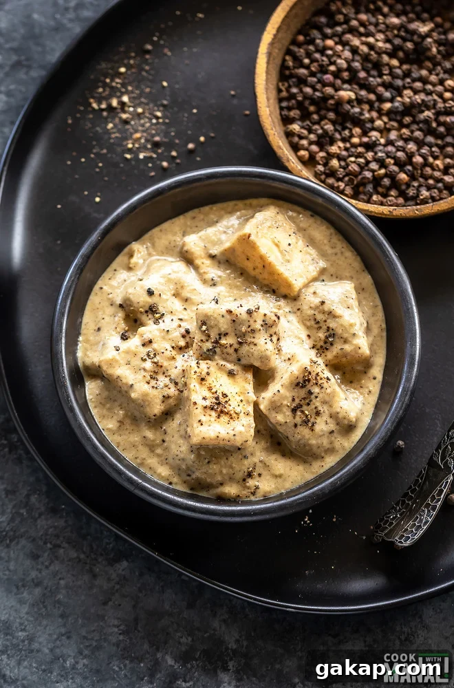 Black Pepper Paneer (Paneer Kali Mirch) — Spicy Indian Cottage Cheese 6 paneer cubes in a white sauce served in a round bowk and a pan of peppercorns placed in the back