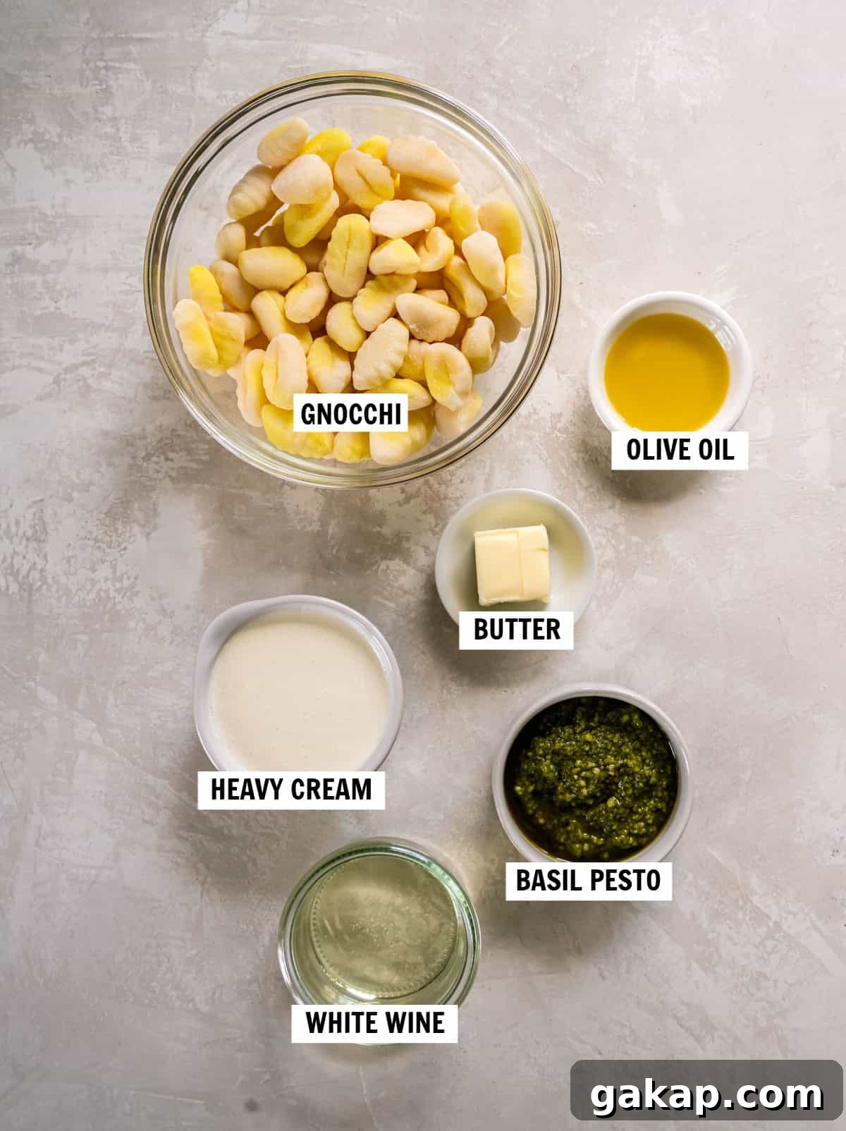 All of the ingredients for pesto gnocchi sitting in bowls on a kitchen countertop including gnocchi, olive oil, butter, heavy cream, basil pesto and white wine.