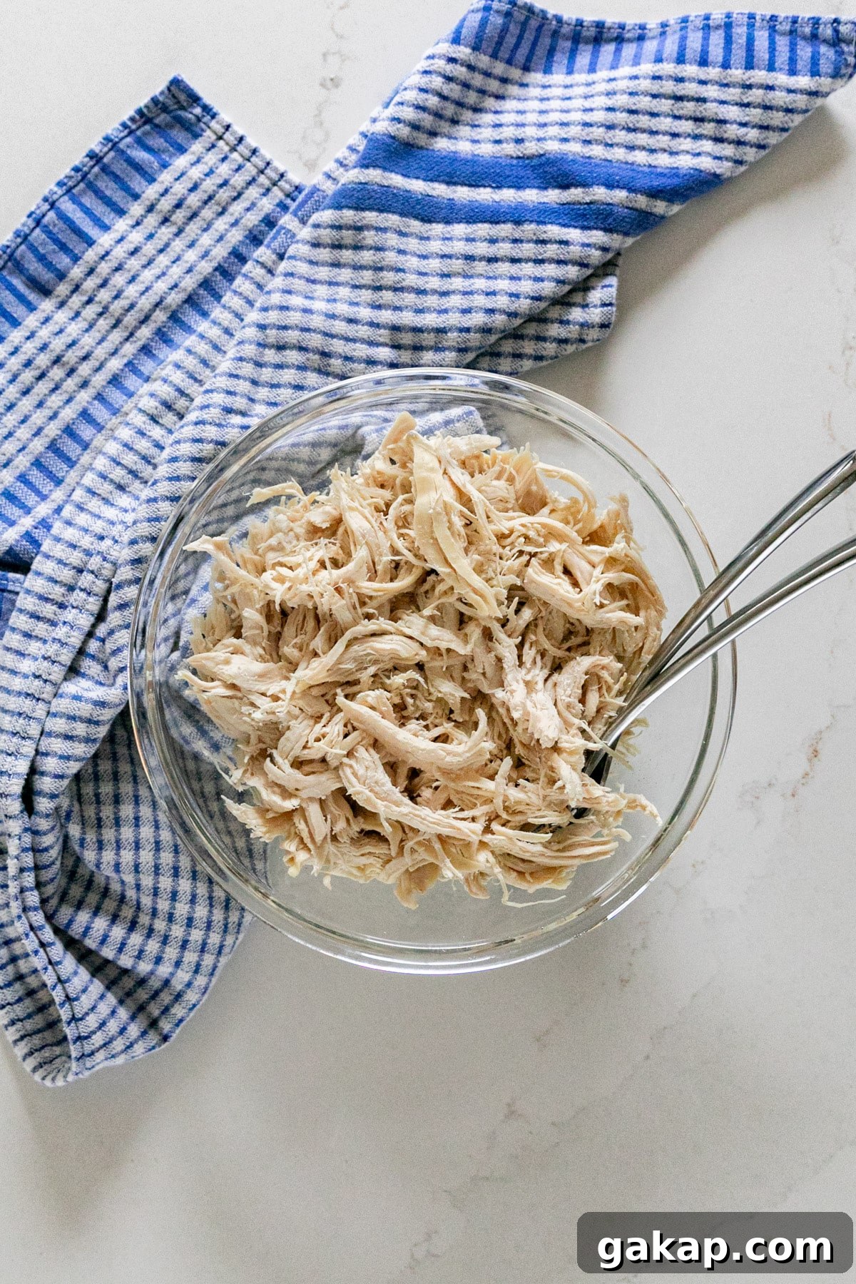 An overhead image of shredded chicken breast in a glass bowl with a blue checked towel next to it and a couple of forks in it.