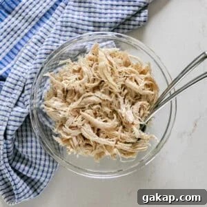 An overhead image of shredded chicken breast in a glass bowl with a blue checked towel next to it and a couple of forks in it.