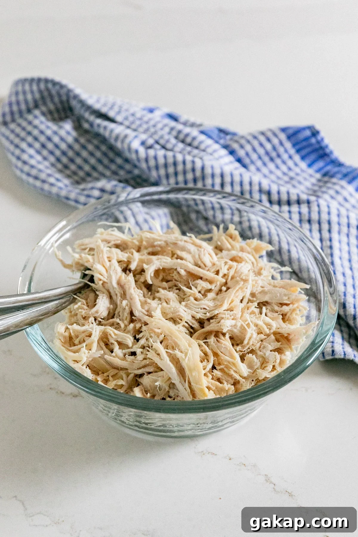 A bowl of shredded chicken with two forks sticking out and a blue checked towel behind it.