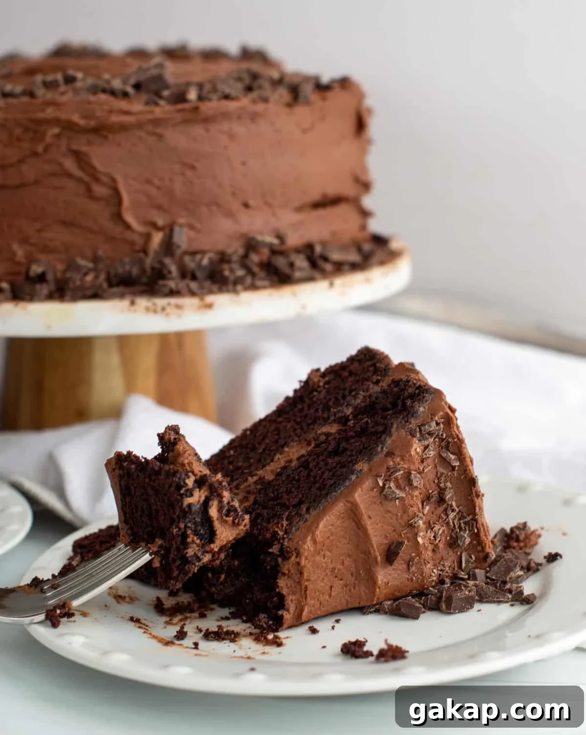 Slice of sourdough chocolate layer cake with chocolate frosting and chocolate shavings on a white plate, with a fork holding a bite and the frosted cake on a stand in the background.