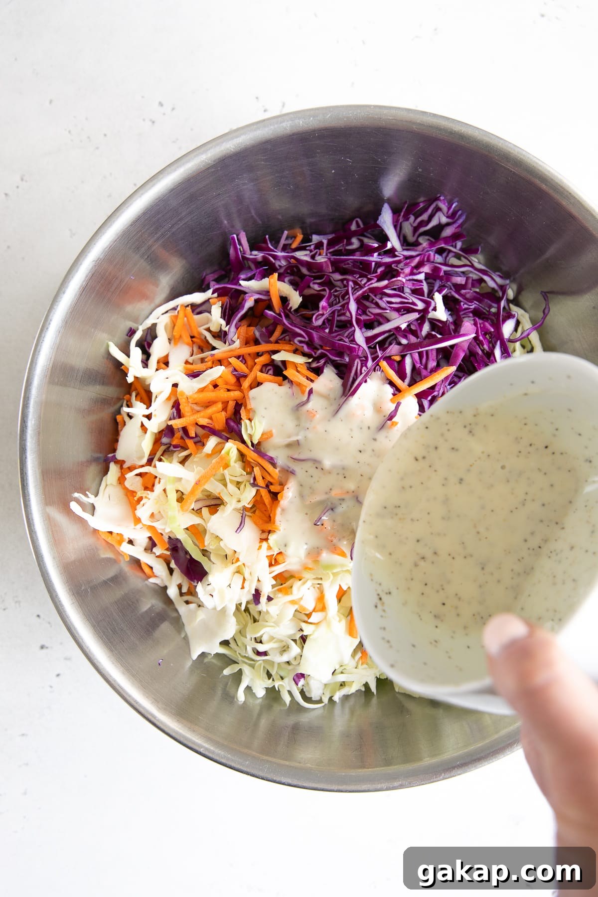 Pouring prepared coleslaw dressing over a bowl filled with shredded cabbage and carrots.