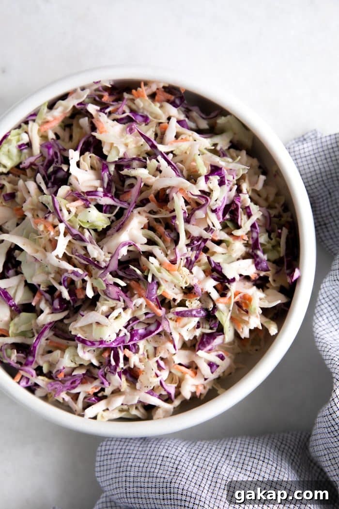 A large white serving bowl filled with freshly made creamy coleslaw, featuring a mix of green and purple cabbage and shredded carrots, dressed in a homemade mayonnaise-based dressing.