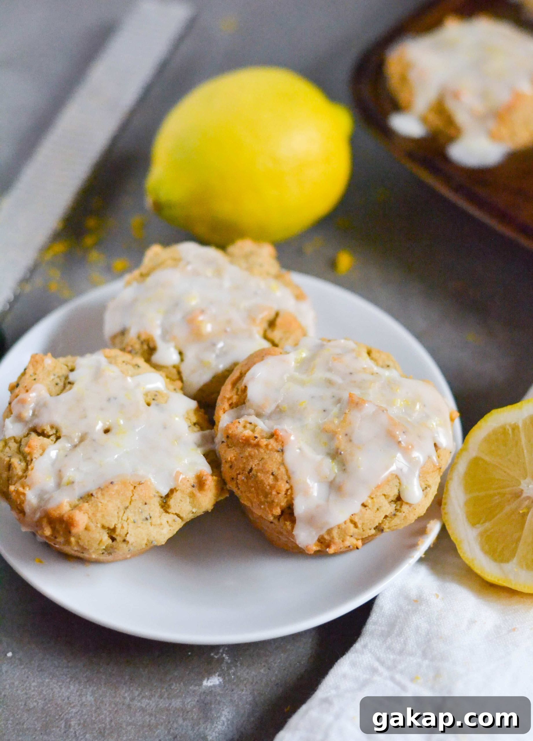 Gluten-Free Vegan Lemon Poppy Seed Muffins Recipe 2 A white plate with three lemon poppyseed muffins with lemon glaze on It with lemons surrounding it.