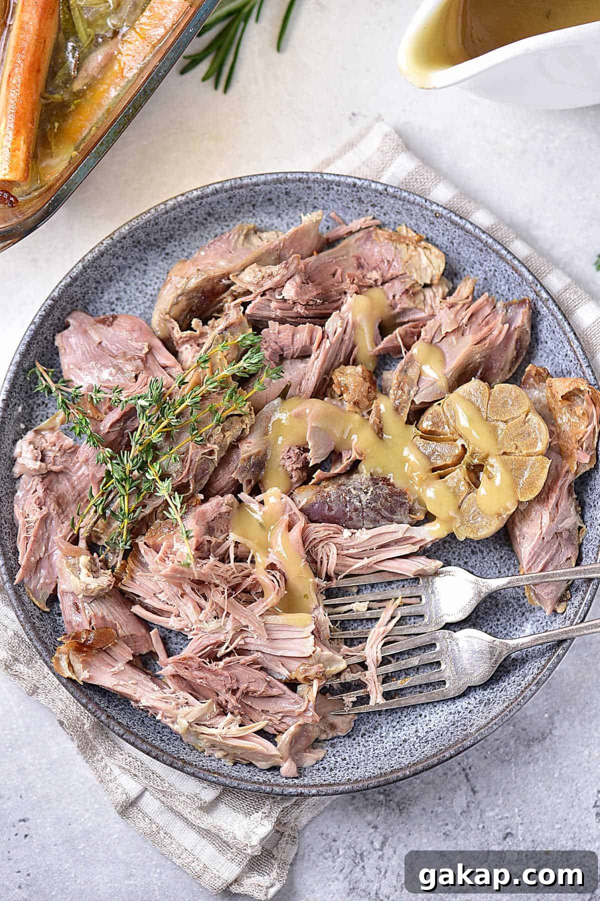 Perfect Slow-Roasted Leg of Lamb Recipe for Tender Meat 11 Serving of pulled-apart leg of lamb with gravy on a grey plate. Silver forks, light grey surface, striped cloth, gravy boat.