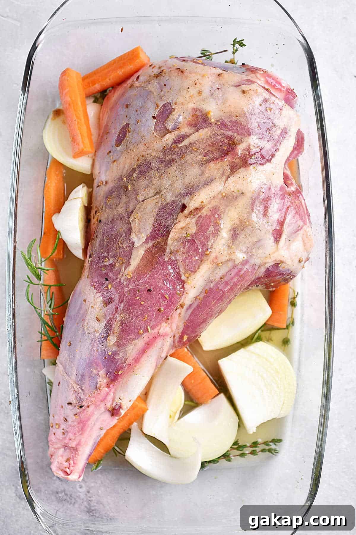 Perfect Slow-Roasted Leg of Lamb Recipe for Tender Meat 5 Glass dish with leg of lamb, herbs, carrots and onions. Light grey surface.