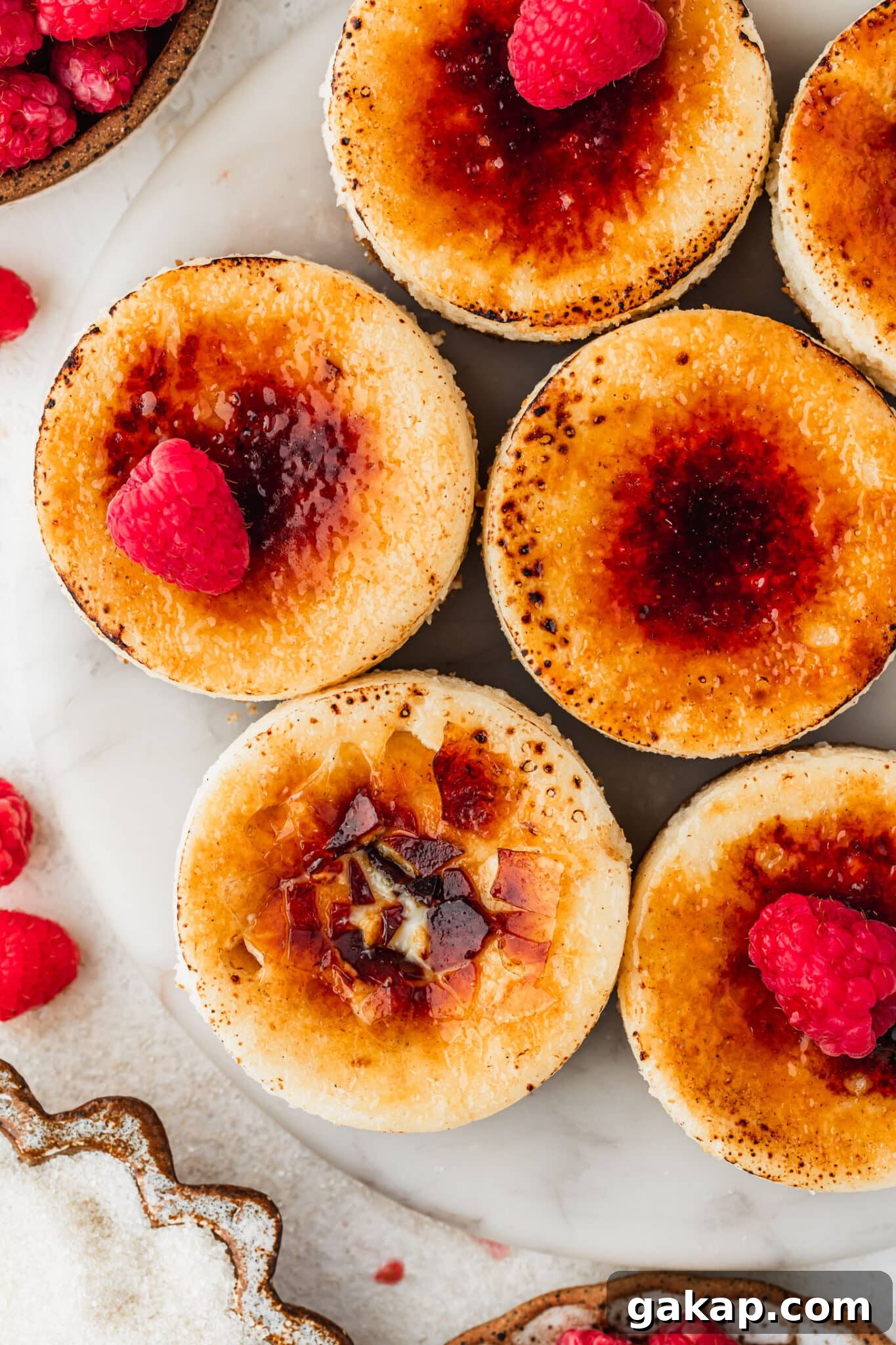 A white platter of mini creme brulee cheesecakes on a white counter next to brown bowls of raspberries and sugar.