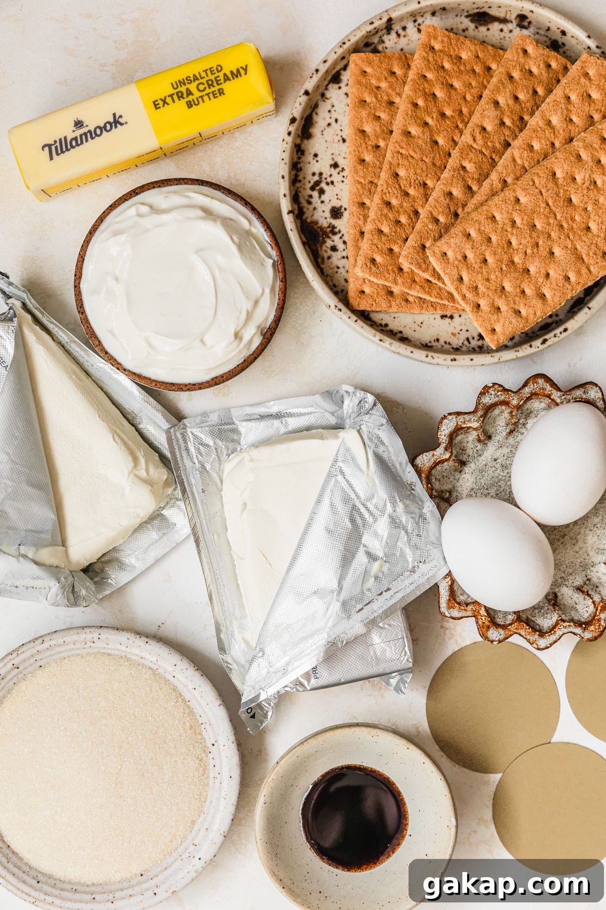 White and brown bowls of graham crackers, sour cream, eggs, sugar, and vanilla bean paste next to bricks of cream cheese and a stick of butter on a tan counter.