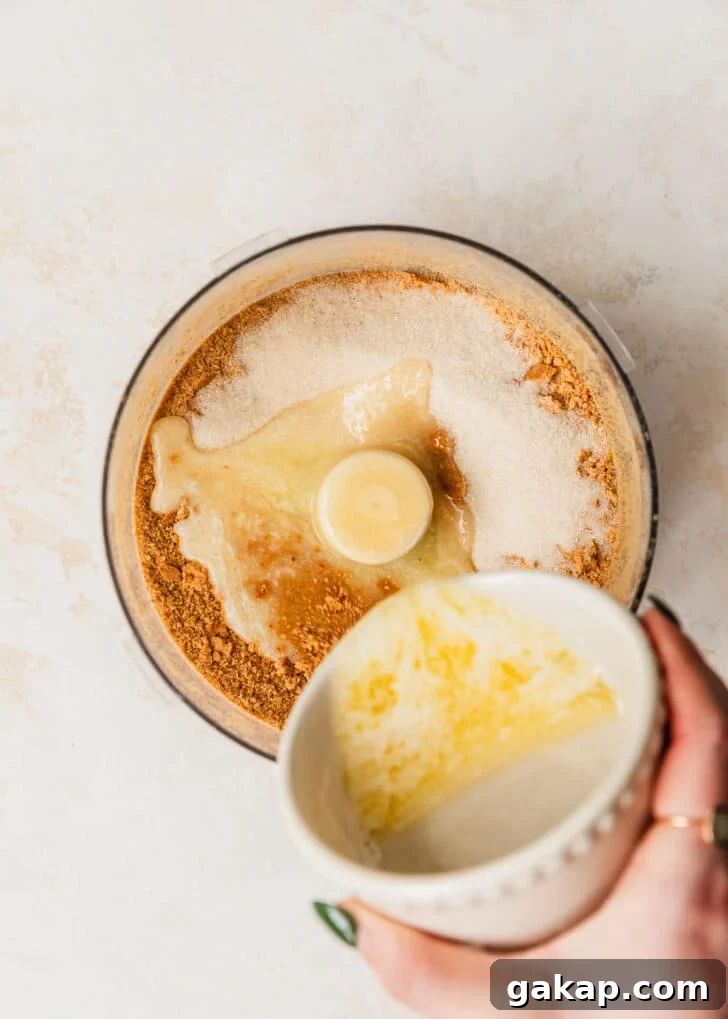 A hand pouring a white bowl of melted butter into a food processor of graham cracker crumbs.