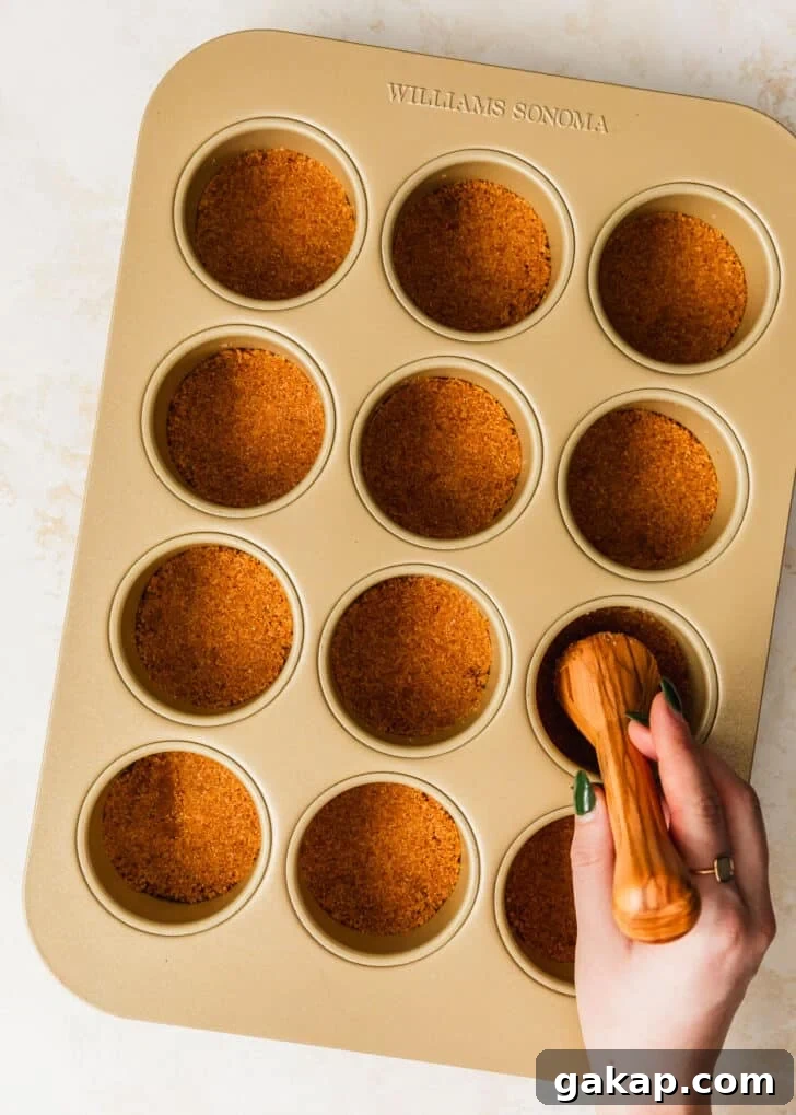 A hand using a pastry tamper to press graham cracker crust into mini cheesecake pans.