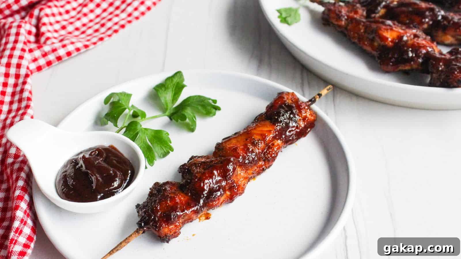 A single barbecued chicken skewer on a white plate, with a small bowl of barbecue sauce and a parsley garnish, next to a plate of additional chicken skewers.