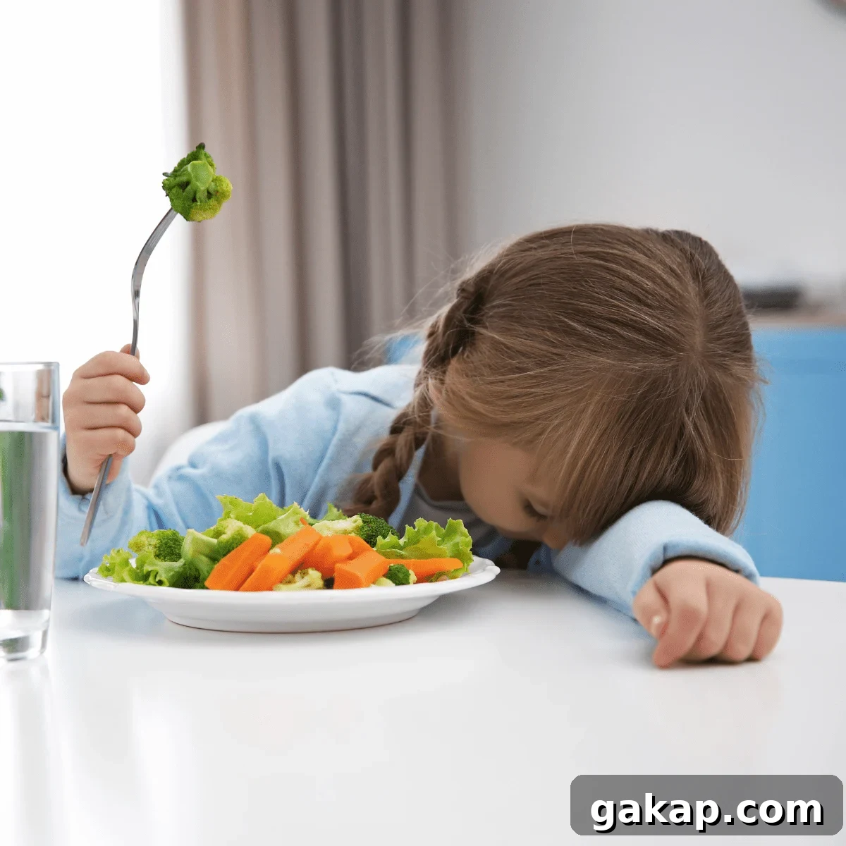 Young girl with head down on table, next to a plate of vegetables, holding a fork in the air with a piece of broccoli on it