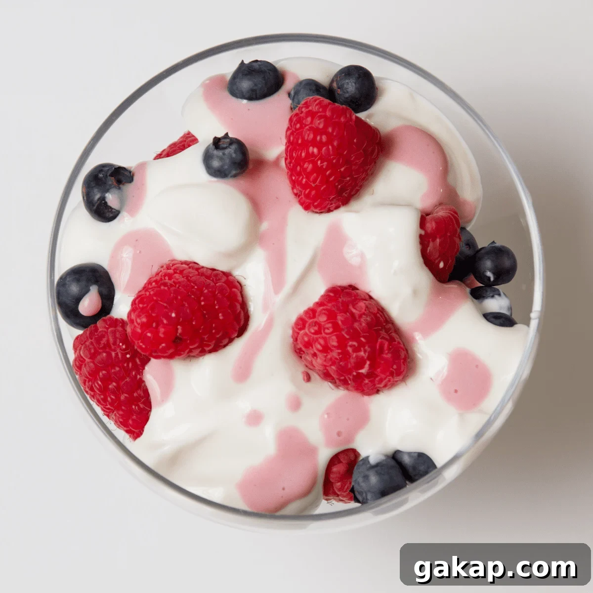 Clear bowl on white table, filled with white yogurt with blueberries, raspberries and pink fruit juice drizzled on top 