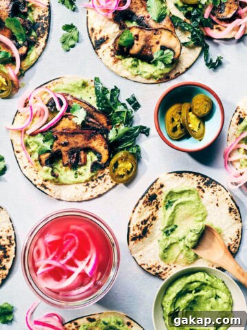 Wide shot of five smoky mushroom tacos on a counter being dressed with toppings like smoky mushrooms, avocado crema, pickled red onions and freshly chopped kale