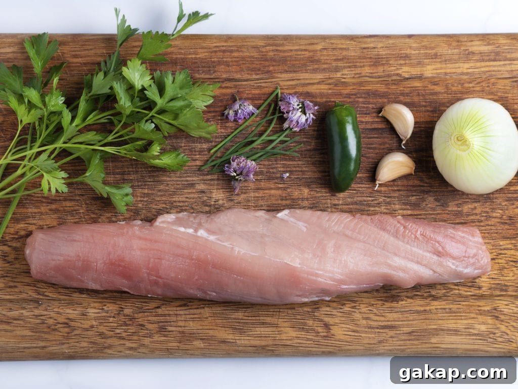 Ingredients on a cutting board: raw pork tenderloin, jalapeno pepper, fresh herbs and garlic