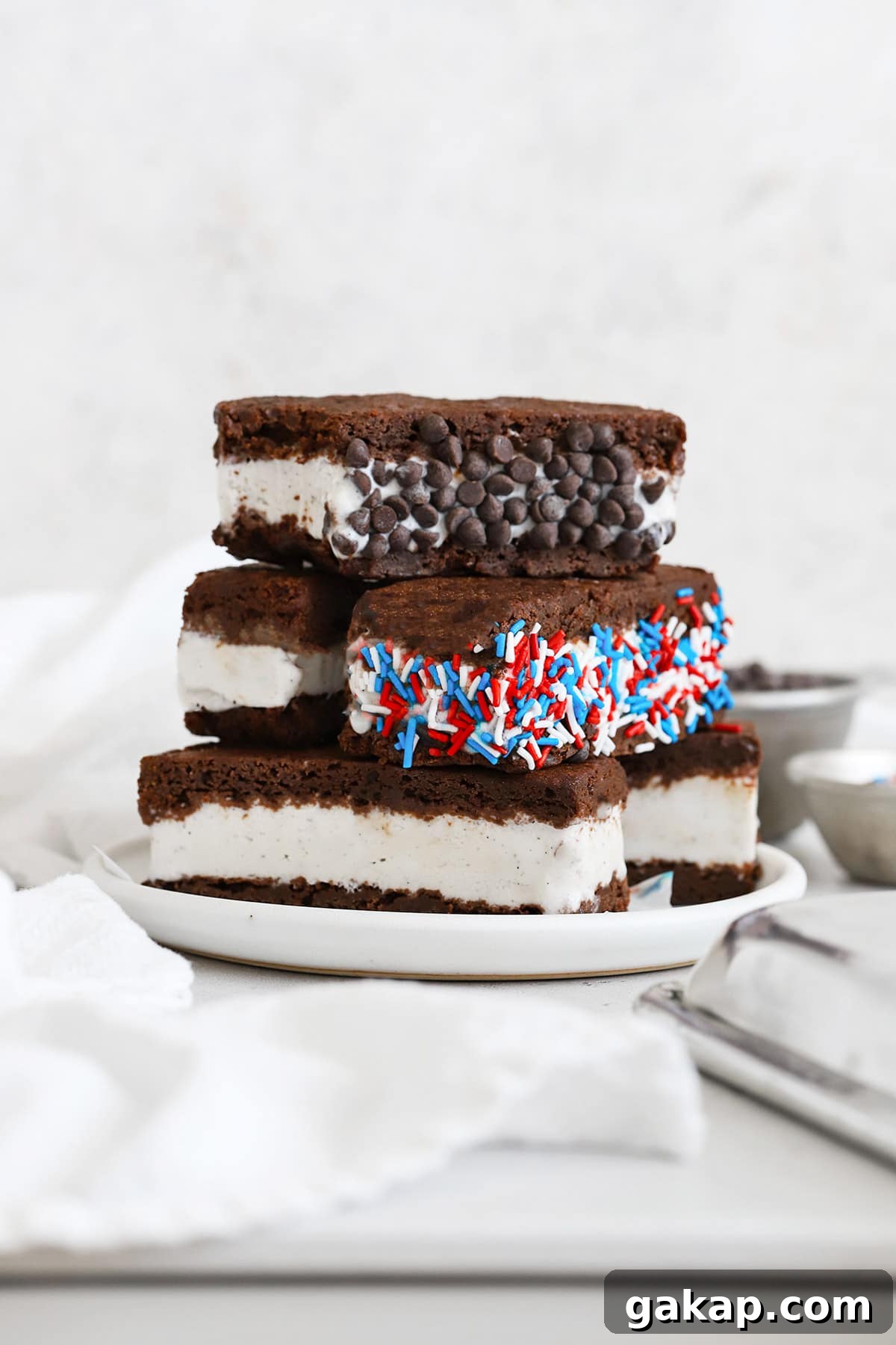 Gluten-Free Brownie Ice Cream Sandwich Recipe 9 Gluten free ice cream sandwiches stacked on a plate. One is rolled in mini chocolate chips and one is rolled in red, white, and blue jimmies sprinkles