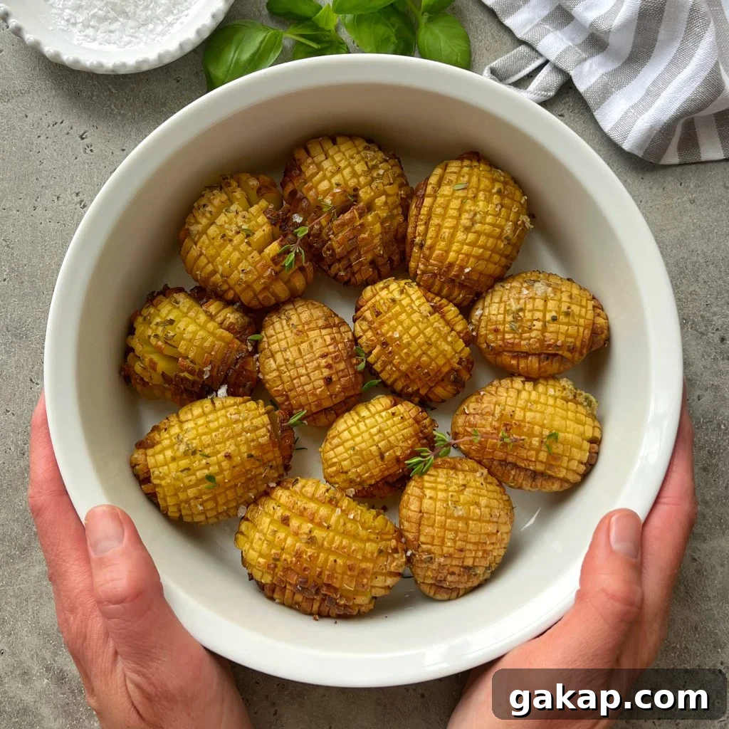 Crispy Hasselback Potatoes with Garlic Butter and Parmesan 2 A pair of hands holds a white bowl filled with twelve hasselback baked potatoes, garnished with herbs. The bowl rests on a gray surface with a striped cloth, fresh basil leaves, and a dish of salt nearby.