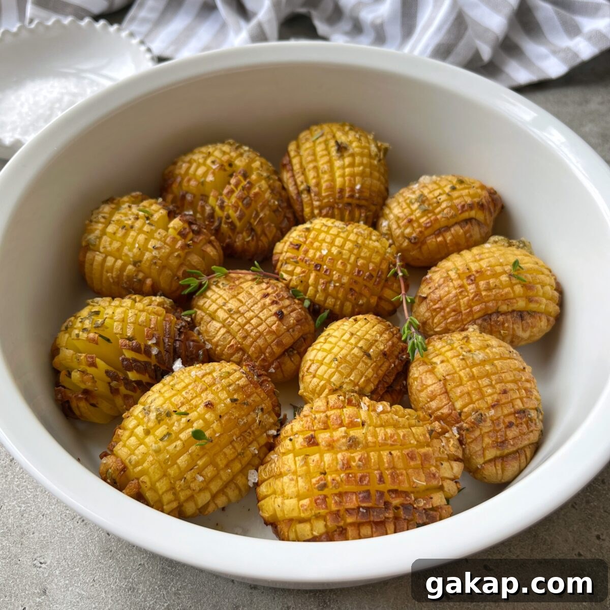 Crispy Hasselback Potatoes with Garlic Butter and Parmesan 3 A white bowl filled with several Hasselback Baked Potatoes, garnished with herbs and coarse salt, sits on a gray surface.