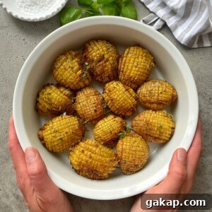Crispy Hasselback Potatoes with Garlic Butter and Parmesan 8 A pair of hands holds a white bowl filled with twelve hasselback baked potatoes, garnished with herbs. The bowl rests on a gray surface with a striped cloth, fresh basil leaves, and a dish of salt nearby.