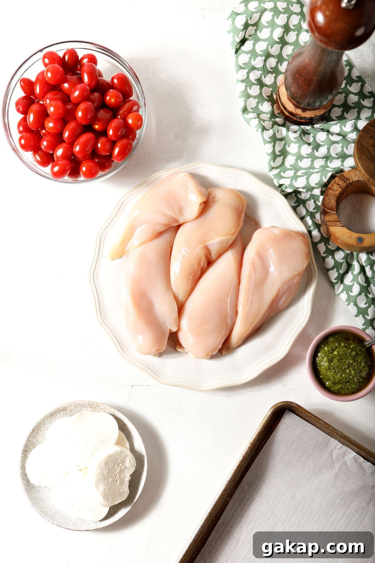 Ingredients to make baked caprese chicken on a white surface next to a green patterned linen