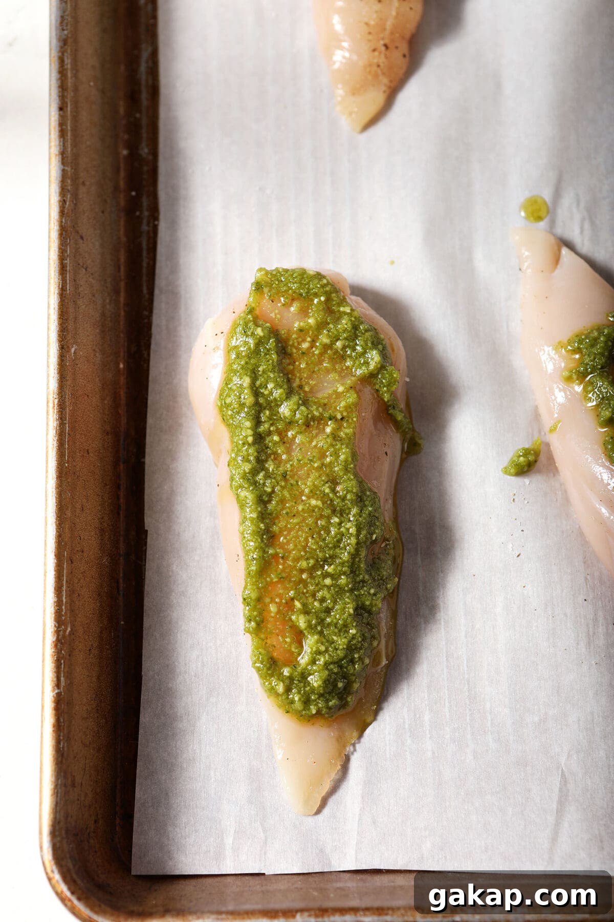 A chicken breast smeared with pesto on a parchment-lined sheet pan
