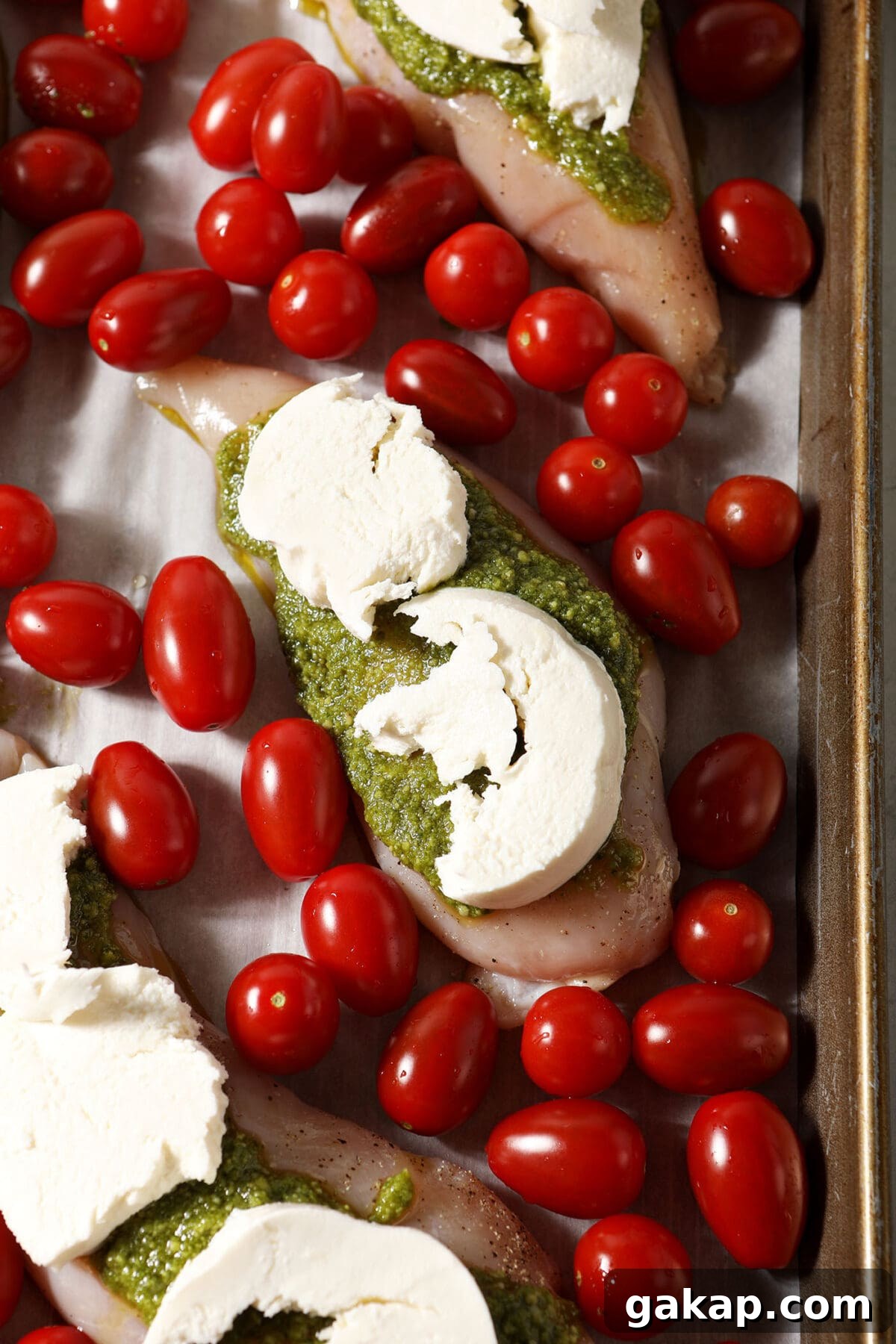 Raw chicken breasts topped with pesto and mozzarella surrounded by cherry tomatoes on a sheet pan