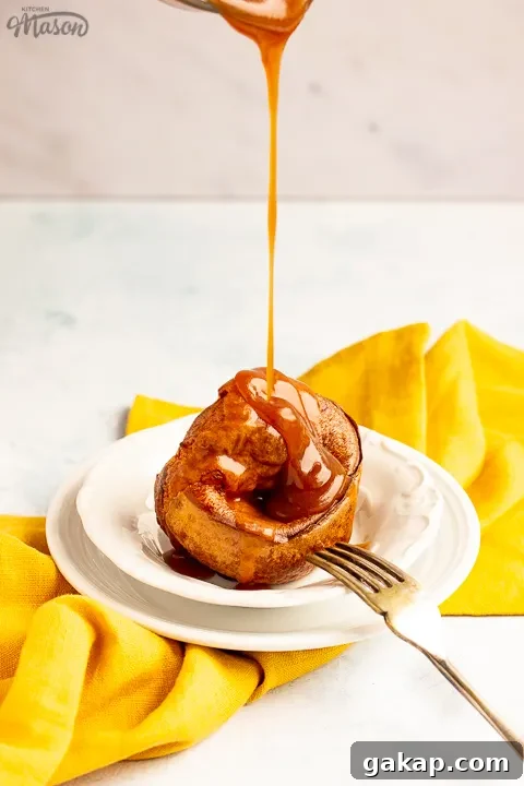 A Yorkshire pudding with gravy being poured over it in a small white bowl set over a mustard yellow napkin. There's a fork resting on the bowl.